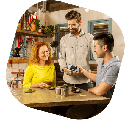 Três pessoas estão em um restaurante, sendo dois clientes e um vendedor. Os clientes são uma mulher branca de cabelo ruivo e expressão sorridente, vestindo uma camiseta amarela de manga comprida, e um homem branco de cabelo preto e expressão sorridente, vestindo uma camisa cinza de manga curta. Os clientes estão pagando a compra aproximando o celular de uma maquininha. O vendedor é um homem branco, de cabelo castanho e expressão sorridente, vestindo uma camisa bege de manga comprida, segurando a maquininha para o cliente aproximar e pagar seu pedido.