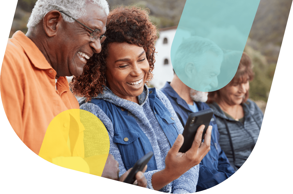 Um homem e uma mulher estão olhando para a tela de um celular. O homem é negro, tem cabelo grisalho e expressão sorridente, está usando um óculos de grau e uma camisa laranja. A mulher é negra, tem cabelos castanhos com mexas claras e expressão sorridente, está vestindo um moletom azul e um colete azul aberto por cima.