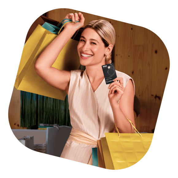 Atriz Luisa Arraes, branca, de cabelo loiro e expressão sorridente, segurando um cartão do PagBank e algumas sacolas de compras. Ela está usando um vestido rosa claro com algumas listras finas e um laço na cintura.