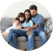 Família composta por uma menina, a mãe e o pai, todos brancos com cabelo preto. A menina está usando um vestido branco, a mãe está usando uma camisa azul clara de manga comprida e uma calça jeans, e o pai uma camisa azul de manga comprida dobrada e uma calça jeans. Todos estão sentados em um sofá cinza e olhando a tela do mesmo celular.
