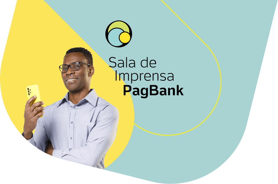 Homem preto sorridente segurando um celular amarelo em frente à parede com a frase 'sala de imprensa PagBank'.