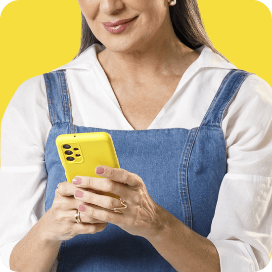 Mulher branca, com cabelo castanho, veste uma jardineira sobre uma camiseta e segura um celular amarelo. A imagem faz referência ao Cofrinho PagBank, produto que ajuda o usuário a guardar dinheiro de forma prática e automática.