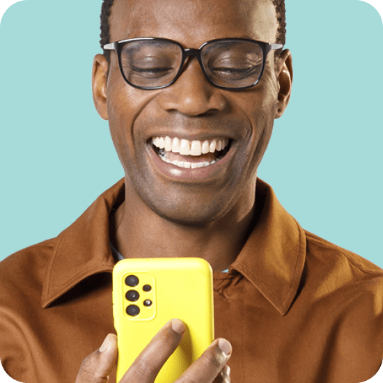 Homem de pele preta, usando óculos e camisa marrom, sorri enquanto segura um smartphone amarelo. A imagem faz referência ao Empréstimo Consignado INSS do PagBank, uma linha de crédito acessível com parcelas descontadas diretamente do benefício ou aposentadoria, contratada de forma prática pelo aplicativo.