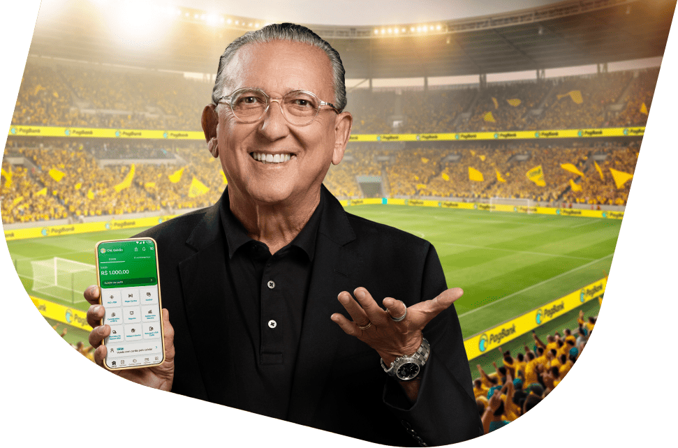 Galvão Bueno segurando smartphone com aplicativo PagBank em estádio de futebol com placas publicitárias do PagBank, símbolo de banco digital completo.