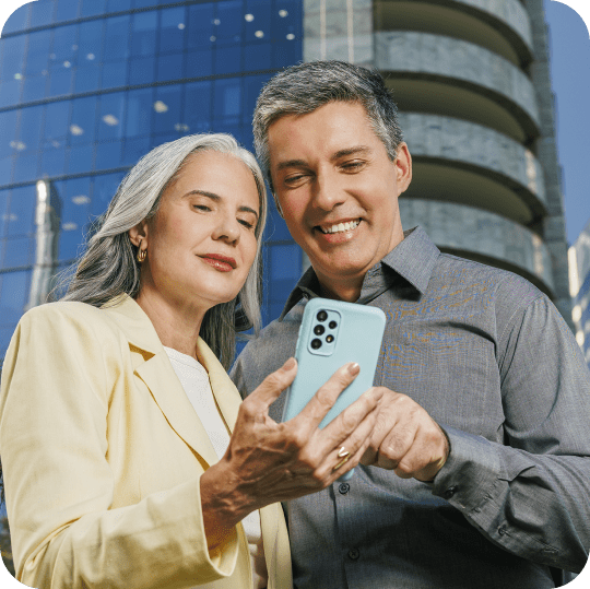 Duas pessoas observam informações em um smartphone em frente a um edifício corporativo, representando o acompanhamento de investimentos e aplicações financeiras.