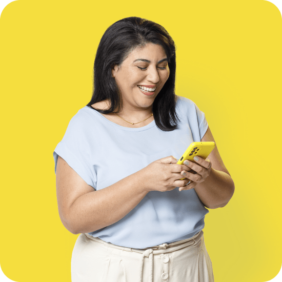 Mulher de pele clara, cabelo escuro e liso, vestindo blusa azul clara e calça bege, segura um celular amarelo e sorri. O fundo é amarelo. A imagem representa o uso do recurso Split de Pagamentos do PagBank, voltado para divisão automatizada de valores entre recebedores em modelos de carrinho único.