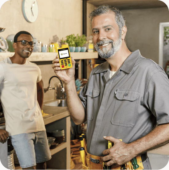 Duas pessoas estão em uma cozinha doméstica. Em primeiro plano, um homem branco vestida com macacão cinza e um cinto de ferramentas, segura uma maquininha de cartão Minizinha NFC 2 PagBAank, voltada para a câmera. Ao fundo, um homem negro encostado na pia, vestindo camiseta branca e shorts jeans. A cozinha possui prateleiras com pratos, utensílios e plantas decorativas.